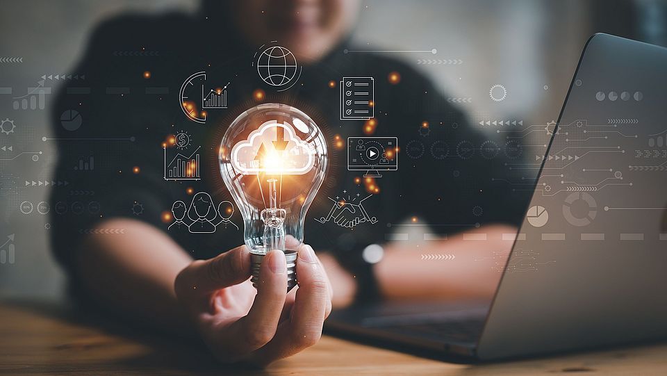 Image of a woman sitting in front of a laptop at a desk, holding in her right hand a luminous light bulb around which circles various symbols symbolizing different theme ideas.