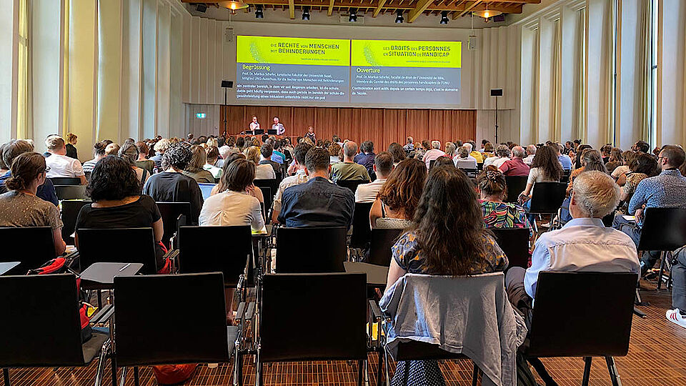 Szene aus der Behindertenrechtstagung 2023, bei der in der Aula der Universität Basel in Reihen sitzende Zuhörer*innen auf die zentral projezierte Fachpräsentation schauen und interessiert zuhören.