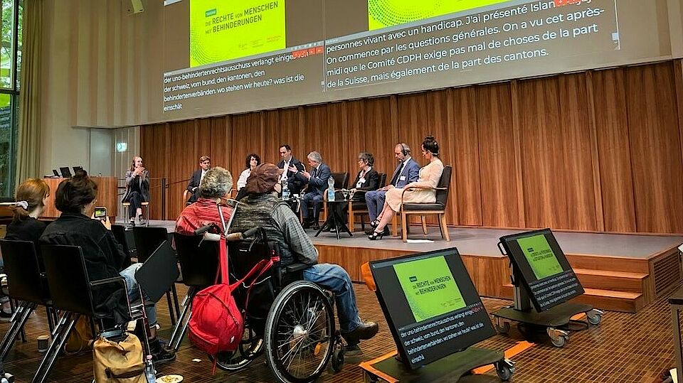 Szenenfoto aus dem Podiumsgespräch-Segment der Behindertenrechtstagung 2022 mit den Podiumsteilnehmenden auf der Bühne und dem aufmerksamen Publikum davor.