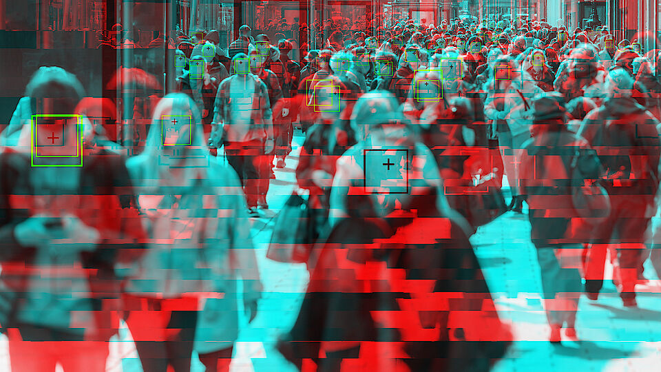 Symbolic image for the topic of facial recognition with a crowd in a pedestrian zone in which individual people have green squares placed around their faces. The whole scene is kept in blue and red colours.
