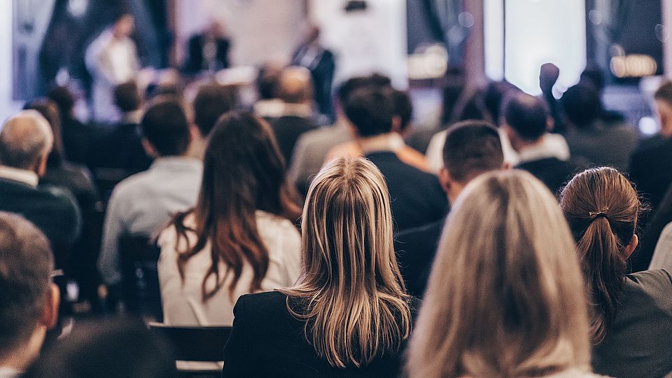 Image looking at the backs of heads of a group of women and men sitting on chairs in a room, looking forward and attentively following an event.