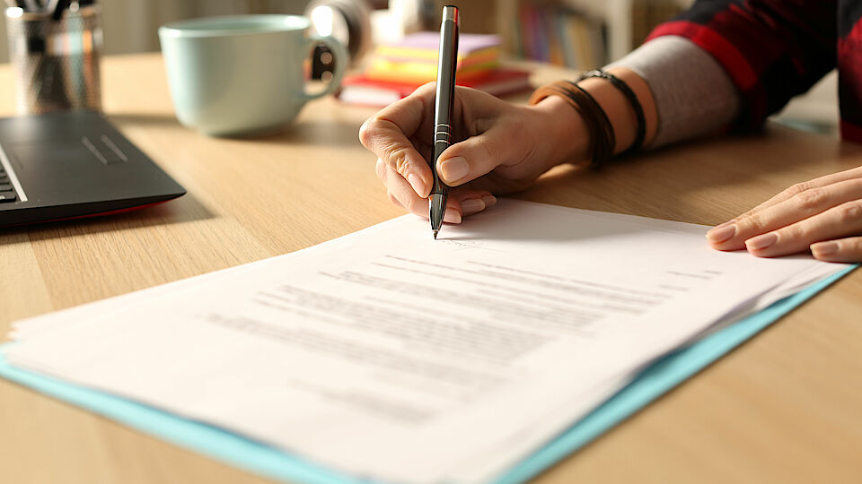Bild eines Papierblattes mit Text, welches auf einem hölzernen Tisch liegt und von einer menschlichen Hand mit einem Kugelschreiber signiert wird.