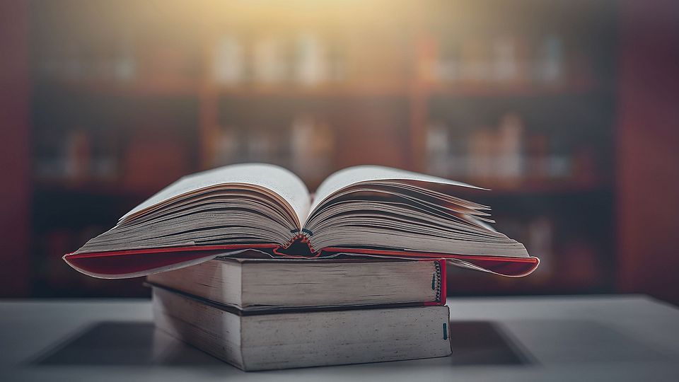 Image of an open book lying on two closed books on a table. In the background there is a blurred library wall with books.