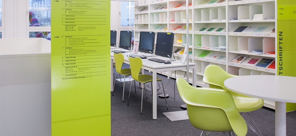 Blick auf Arbeitsplätze in der Bibliothek mit einem Besprechungstisch im Vordergrund und PC-Arbeitsplätzen im Hintergrund. 