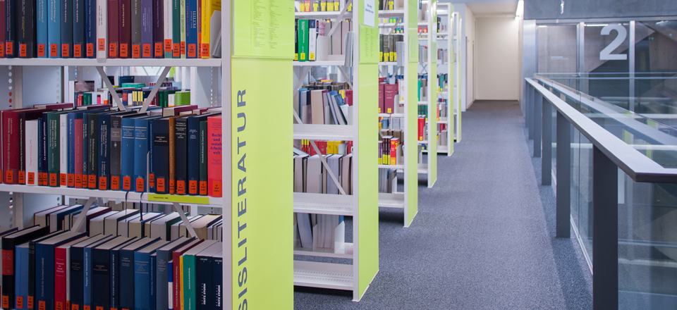 Aufnahme eines Ganges auf dem Bibliotheksstockwerk mit Perspektive auf ein gelbes mit Büchern vollgestelltes Regal.
